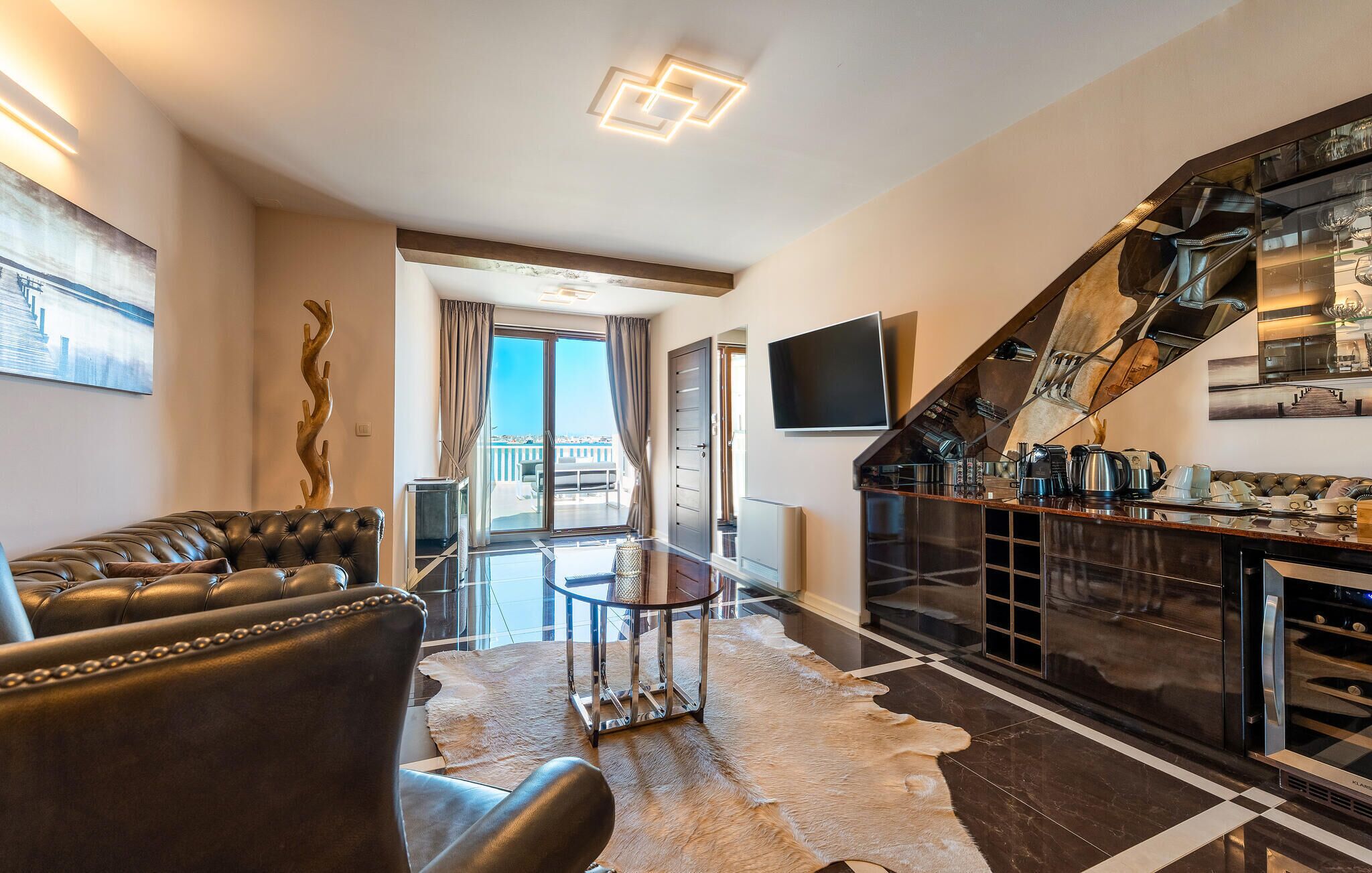 A modern living room with a leather sofa, a glass coffee table, and a fur rug. There is a kitchen area with a stainless steel refrigerator and a dining table.