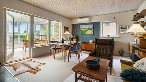 En rummelig stue med store glasdøre til et udendørs område, et spisebord med stole, en læderlænestol og et bogreol.