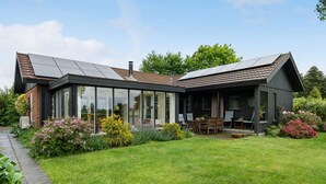 Et moderne hus med store glasfacader, en terrasse med udendørs møbler og en have med forskellige planter.