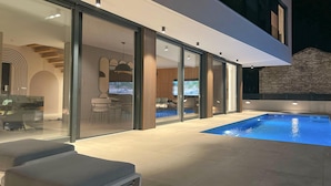 A modern outdoor pool area with a glass-enclosed patio, a stone wall, and a seating area with cushions.