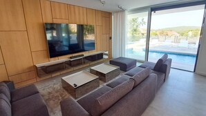 A modern living room with a large flat-screen TV, a sofa, and a coffee table. There is a sliding glass door leading to a pool area.