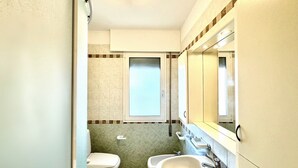 A bathroom with a toilet, bidet, and sink, with a window above the toilet and a mirror above the sink. - Aleksandra 4-Carraro Immobiliare Jesolo (Lido di Jesolo)