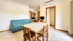 A small, well-lit room with a wooden dining table and chairs, a blue sofa, and a television. - Aleksandra 4-Carraro Immobiliare Jesolo (Lido di Jesolo)
