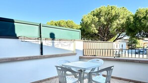 A balcony with a white table and chairs, a green awning, and a tree in the background. - Aleksandra 4-Carraro Immobiliare Jesolo (Lido di Jesolo)