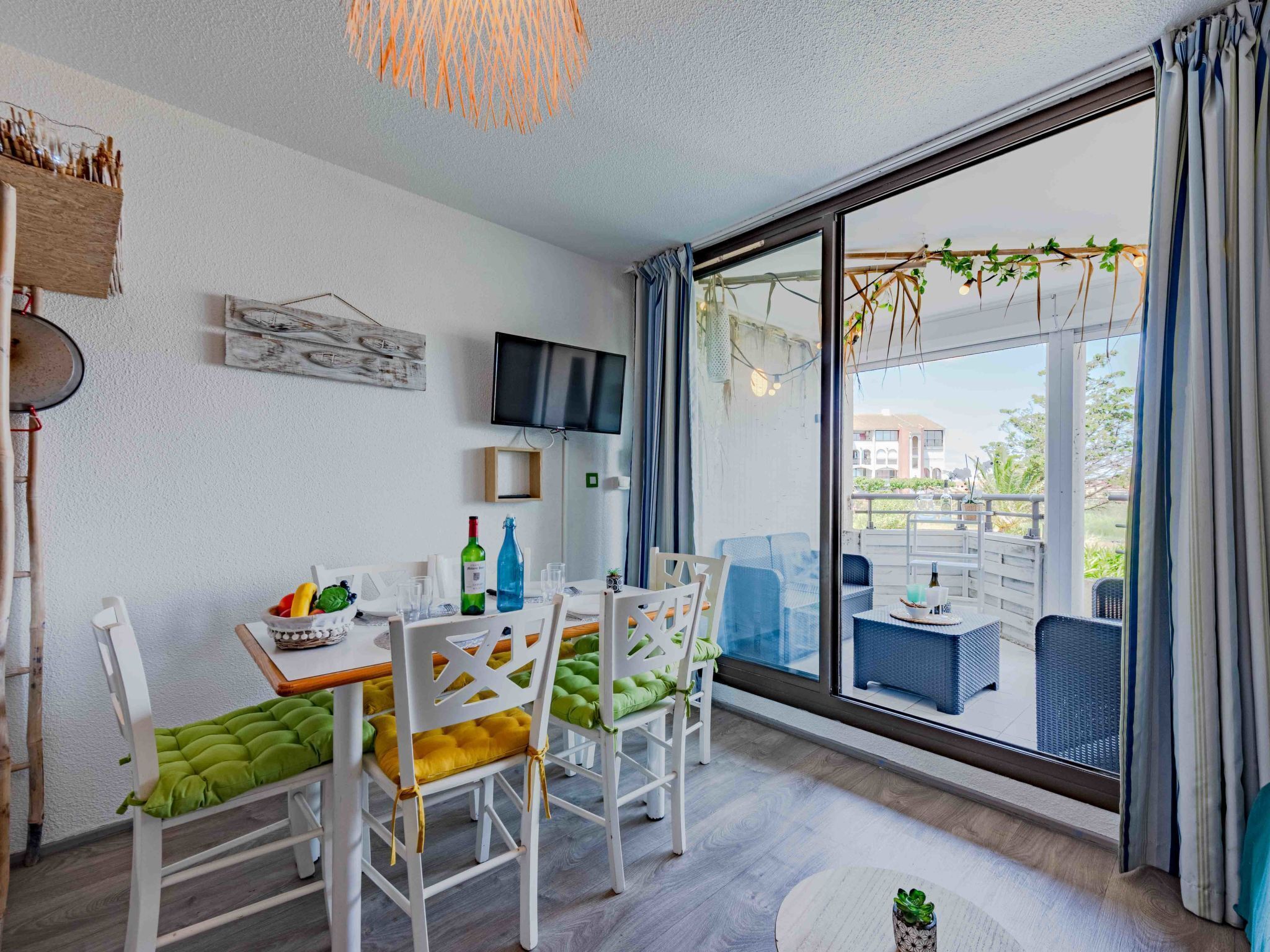Un comedor con una mesa dispuesta para cuatro personas, sillas con cojines, un televisor en la pared y vistas al balcón exterior.