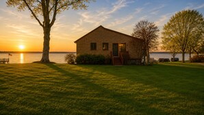 Une cabane au bord d’un lac, au coucher du soleil, avec une pelouse verdoyante, une terrasse en bois et un escalier menant à la porte.