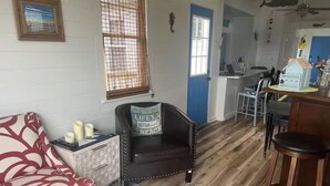 A cozy living room with a patterned sofa, a dark leather chair with a cushion, and a small side table with candles. A kitchen area is visible in the background with a blue door and a window with blinds.