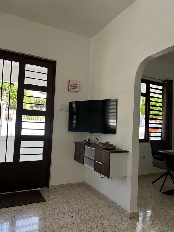 A room with a flat-screen TV mounted on a wall, a wooden desk with drawers, a black chair, and a door with frosted glass panels.