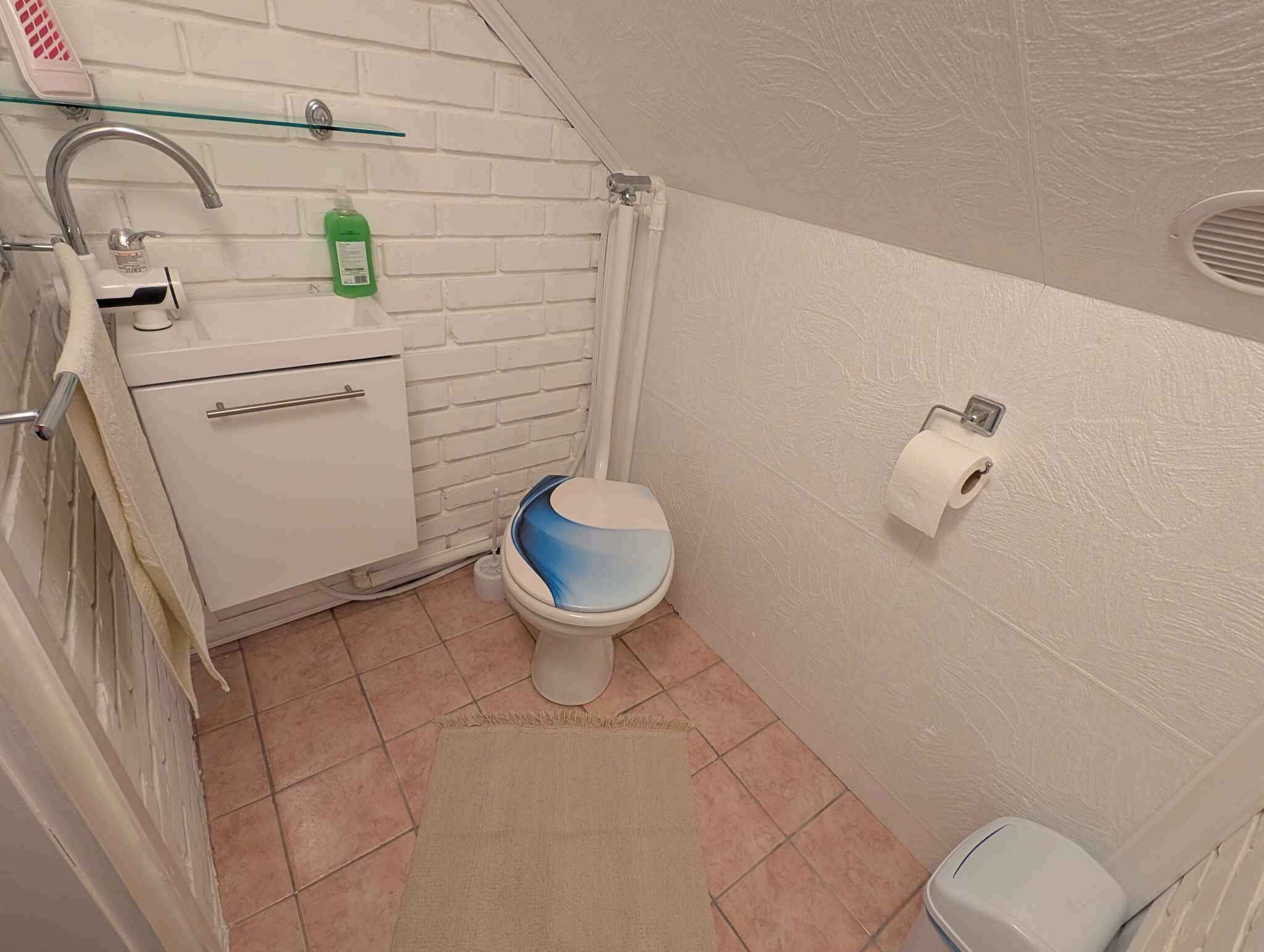 A small bathroom with a white sink, a glass shelf, a toilet, and a white trash bin.