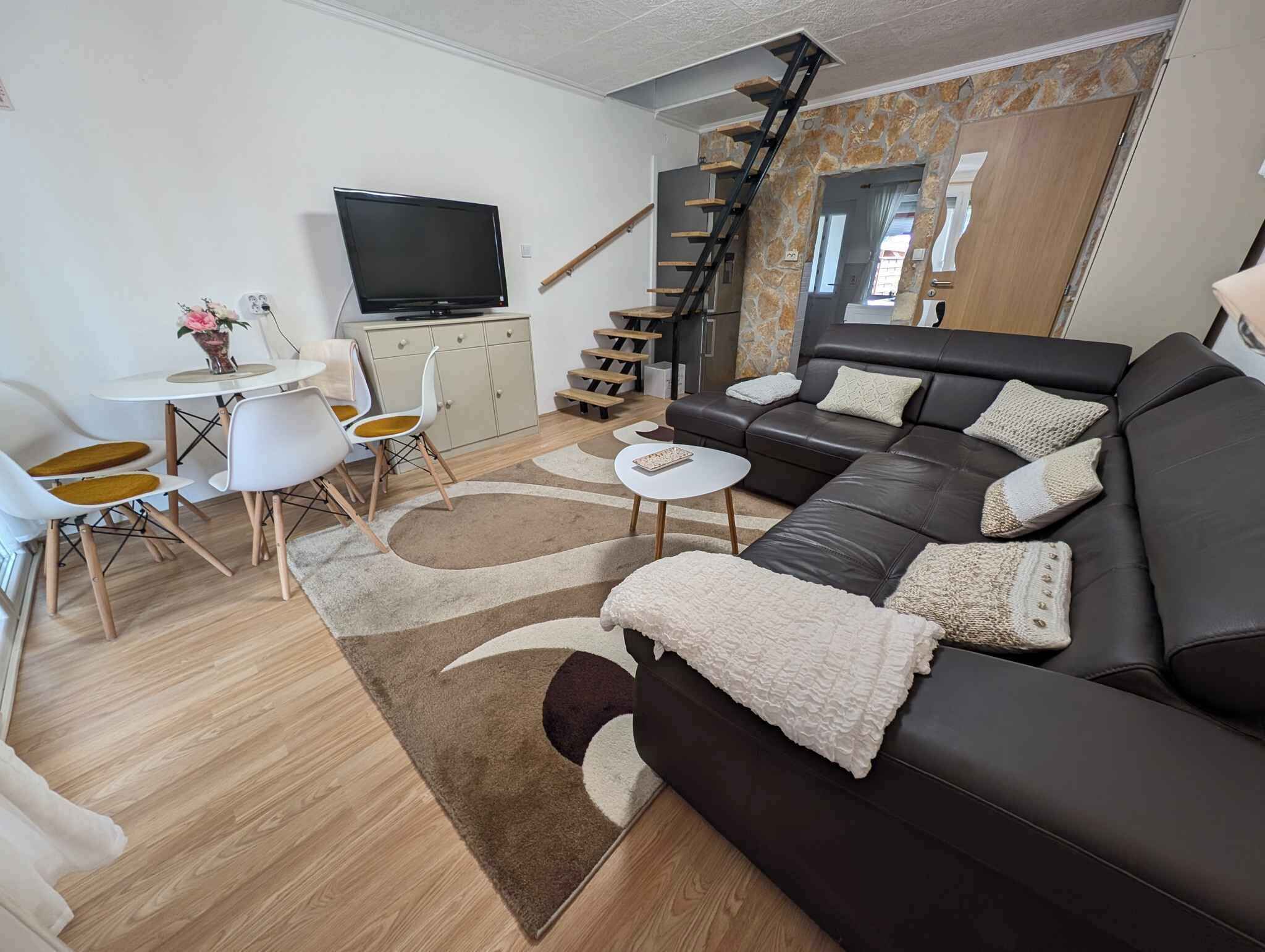 Una sala de estar moderna con un sof á de cuero negro, una mesa de comedor blanca con sillas, un televisor de pantalla plana y una escalera que lleva a un nivel superior.