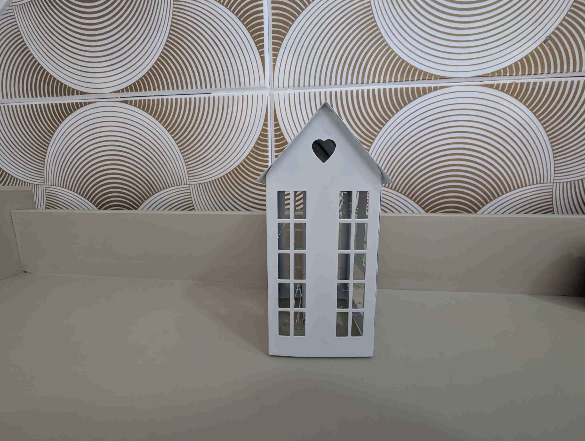 A white house-shaped object with a heart on top, placed on a beige surface against a patterned wall.