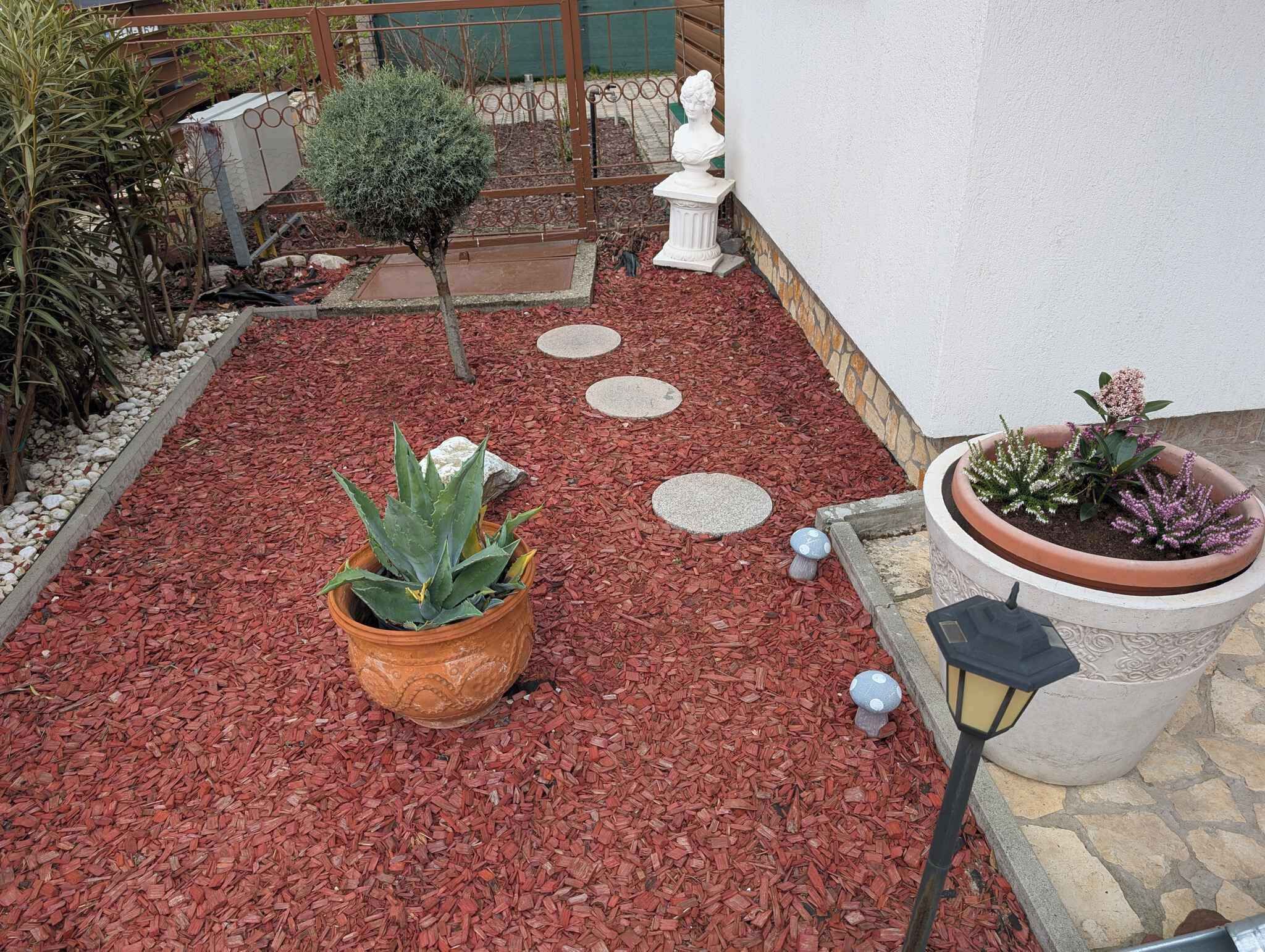 Ein Garten mit roter Mulchschicht, Topfpflanzen, einem weißen Büstenporträt und trittfesten Steinen.