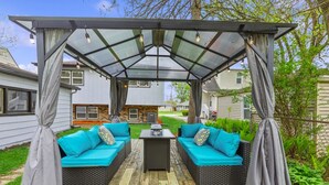 Un porche cubierto con dos sofás y una mesa, rodeado por una pérgola con cortinas.