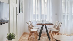Un comedor con una mesa de madera dispuesta para cuatro personas, rodeada de sillas, en una habitación con paredes blancas y ventanas amplias con cortinas translúcidas.