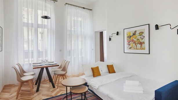 Una habitación de hotel moderna con una cama, una mesita redonda pequeña, dos sillas, un tapete colorido y un ventanal con cortinas translúcidas.