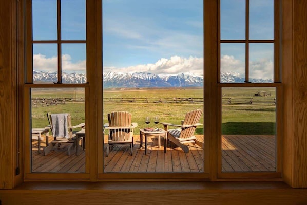 A room with a large window overlooking a snowy mountain range, wooden deck, and two Adirondack chairs.