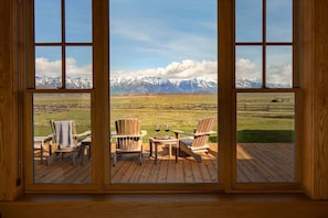 Habitación con amplia ventana que da a una cadena montañosa nevada, terraza de madera y dos sillas Adirondack.