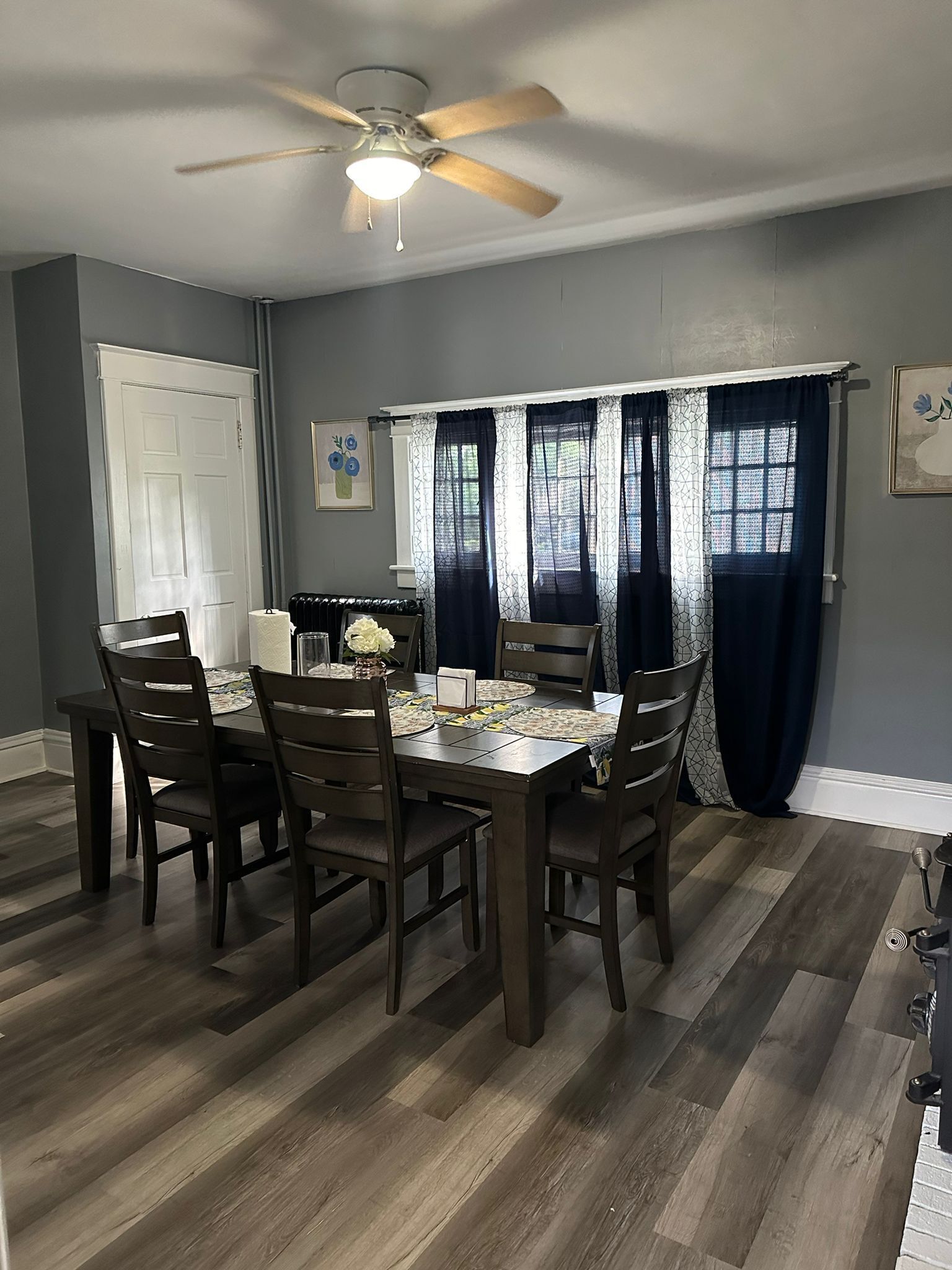 A dining area with a table set for four, surrounded by chairs, a ceiling fan, and a window with patterned curtains.