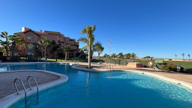 A swimming pool with a clear blue water, surrounded by palm trees and a building in the background. - Casa Serenidad Mar Menor Golf Resort (Torre Pacheco)