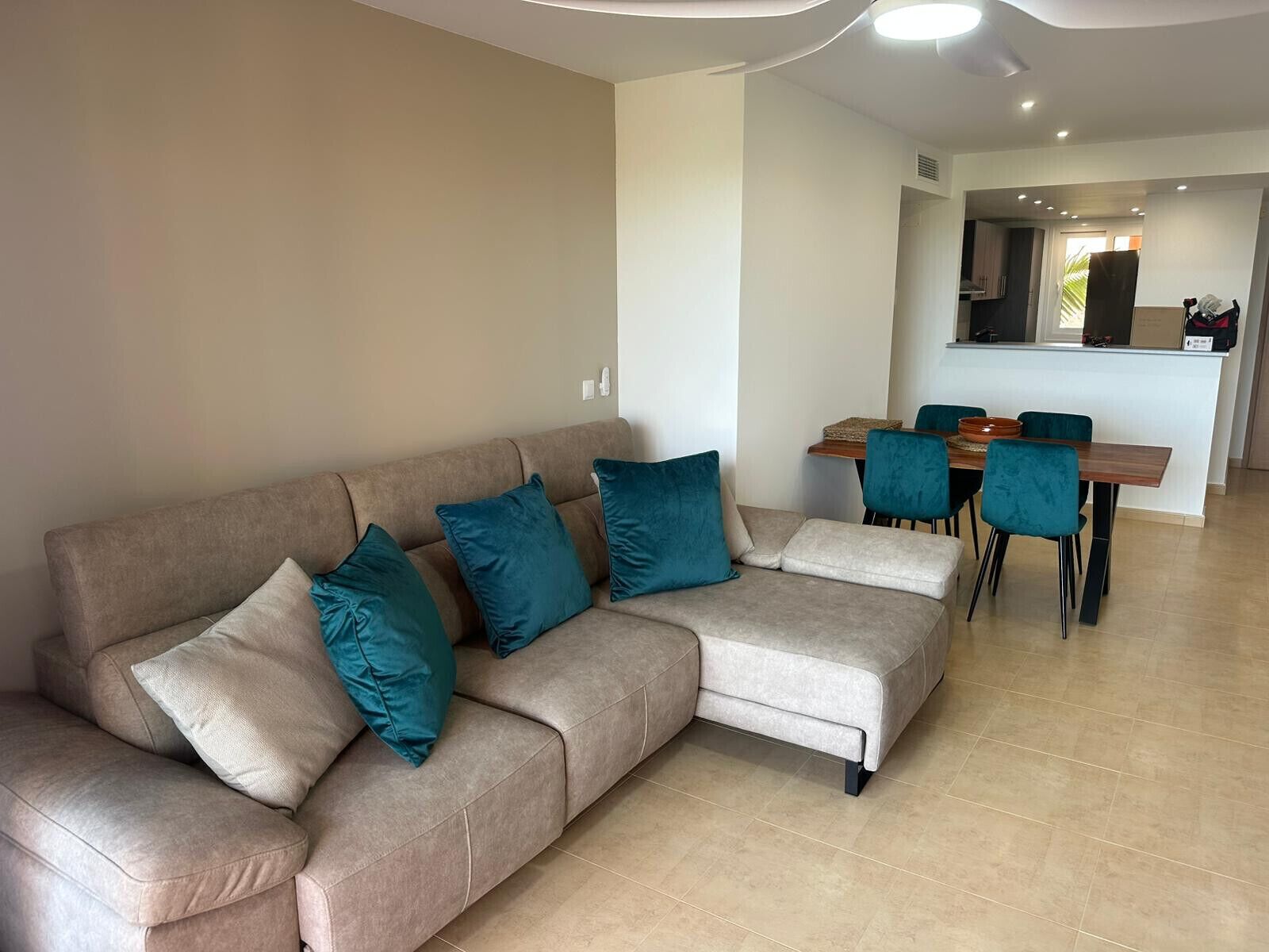 Een moderne woonkamer met een beige modulaire bank, teal kussens en een eethoek met een houten tafel en teal stoelen.