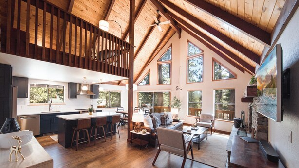 A spacious loft with a high ceiling, wooden beams, a kitchen with a central island, a living area with a fireplace, and large windows offering views of the outdoors. - Hillcrest at Tahoe Donner by Tahoe Getaways - 3BR Pet-Friendly w/ Pool Table & Ping Pong (Truckee)