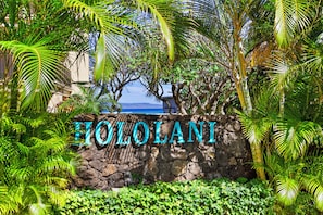 Um muro de pedra com o nome "HILO LANI" em letras grandes, cercado por folhagem tropical exuberante.