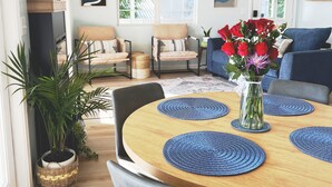 A dining area with a round wooden table, blue placemats, and a vase of red flowers. There are chairs around the table, and a sofa is visible in the background.