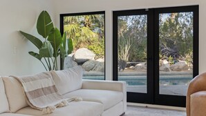 A modern living room with a white sofa, a large plant, and a view of a pool through glass doors. - Palm Springs Haven / Pool, Hot Tub & Mountain Views (Palm Springs)