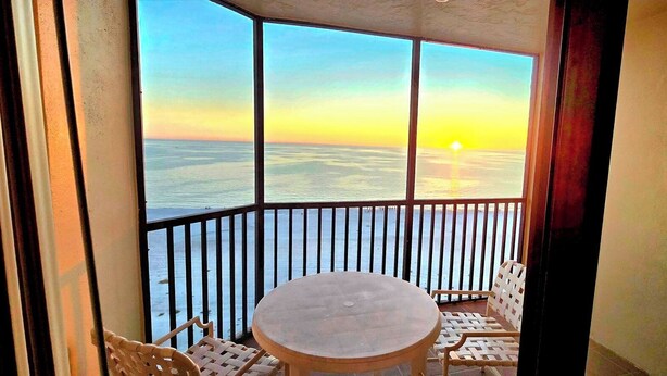 A balcony with a view of the ocean at sunset, a round table, and two wicker chairs.