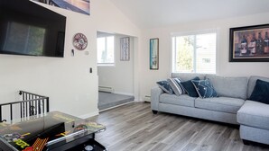 A living room with a grey sofa, a flat-screen TV mounted on the wall, and a foosball table. - Charming 3-bedroom house in Conesus with lake access and dock for boat (Conesus)