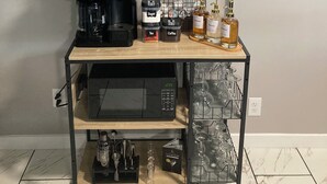 A coffee station with a coffee maker, sugar, tea, and coffee containers, a shelf with mugs and a potted plant, and a rack with glasses.