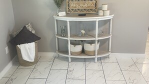 A living room with a flat-screen TV mounted on the wall, a white console table with decorative items, a trash can with a white cloth, and a patterned sofa.
