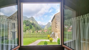 A room with a large window offering a view of a mountainous landscape, a wooden fence, and a pathway leading to a building with a red roof.