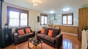 A living room with two sofas, a coffee table, and a fireplace. There is a kitchen area with wooden cabinets and a window.