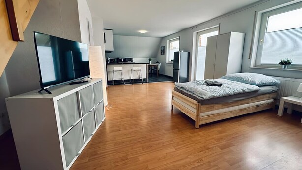 Une chambre moderne avec un plancher en bois, un lit, une télévision et, au fond, un coin cuisine.