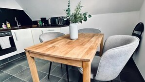 Un coin repas avec une table en bois et deux chaises grises, un vase blanc avec une plante, et une cuisine en arrière-plan.