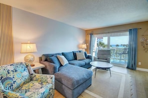 Un salon comprenant un canapé, un fauteuil et une petite table. Il y a un balcon avec vue sur l’océan.