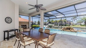 Un vaste espace extérieur aménagé pour les repas, avec vue sur la piscine, un ventilateur de plafond et une horloge murale.
