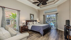 Une chambre à coucher avec un lit, des tables de chevet, un bureau et une chaise. On y trouve un ventilateur de plafond, une fenêtre avec des rideaux et une vue sur une piscine.