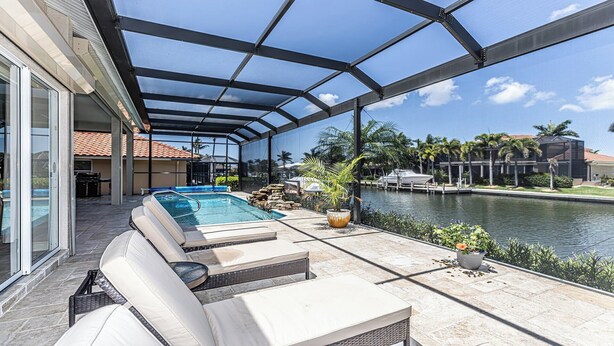 A covered outdoor pool area with lounge chairs, a view of a water body, and a modern building in the background.