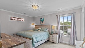 A bedroom with a bed, bedside tables, a nightstand, a chandelier, a window with curtains, and artwork on the walls. - Discover Peaceful Palms, a charming 3-bedroom home in Oak Island's East Beach (Oak Island)
