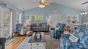 A spacious living room with a ceiling fan, a grey sofa, a blue patterned armchair, and a dining area with a white table and chairs. - Discover Peaceful Palms, a charming 3-bedroom home in Oak Island's East Beach (Oak Island)