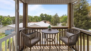 A covered balcony with two wicker chairs and a small table, overlooking a residential area with trees and houses. - Discover Peaceful Palms, a charming 3-bedroom home in Oak Island's East Beach (Oak Island)