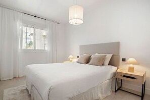 A modern bedroom with a large bed, two bedside tables with lamps, a ceiling light, and a view through a window with curtains.