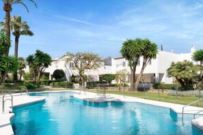 A swimming pool with a clear blue water, surrounded by palm trees and a white building with a tiled roof.