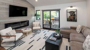 A modern living room with a flat-screen TV, a fireplace, a sofa, a coffee table, and a large window with a view of the outdoors.