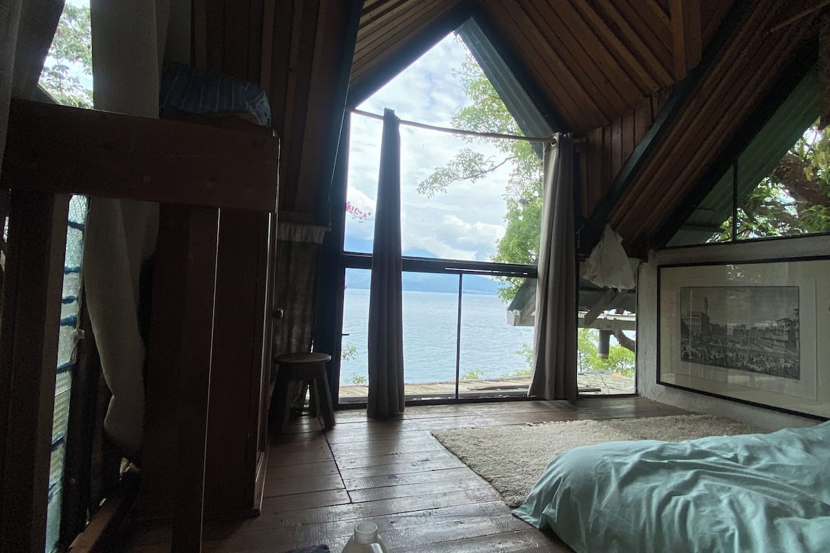 Een kamer met houten vloer, een bed, een tafel met een kopje erop en uitzicht op zee door grote ramen.