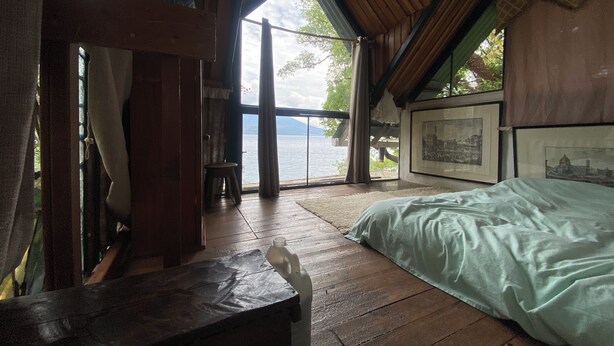 A room with a bed, wooden floor, a bench, a window with a view of a body of water, and framed pictures on the wall. - Blue Forest (San Marcos La Laguna)