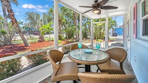 A covered outdoor seating area with wicker chairs and a glass table, overlooking a landscaped garden and palm trees. - Low Tide – Pool, Pet Friendly, In SK Village - Walk Everywhere! (Siesta Key)