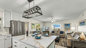 A modern kitchen with white cabinets, a stainless steel refrigerator, and a marble island. There is a dining area with a table and chairs, and a living room with a sofa and a lamp. - Low Tide – Pool, Pet Friendly, In SK Village - Walk Everywhere! (Siesta Key)
